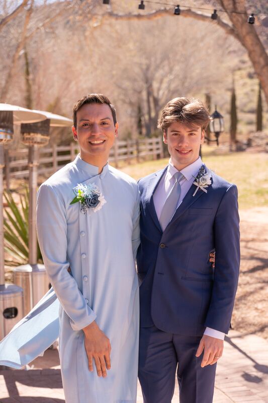 Two grooms with boutonnieres smiling outdoors at a wedding